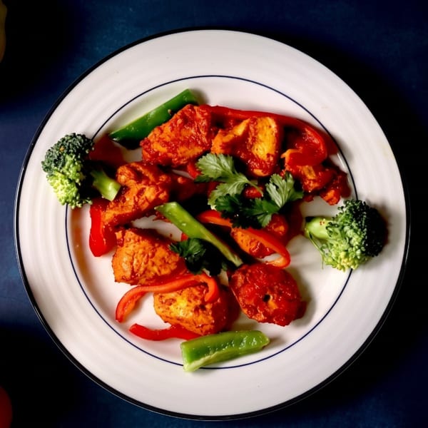 Spicy chicken pieces with red and green bell peppers, broccoli, and cilantro on a white plate.