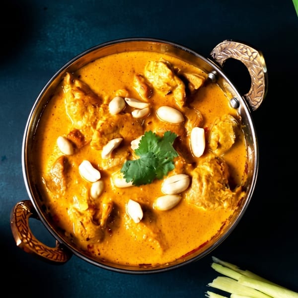 Golden chicken curry with peanuts and cilantro in a brass bowl on a dark blue surface.